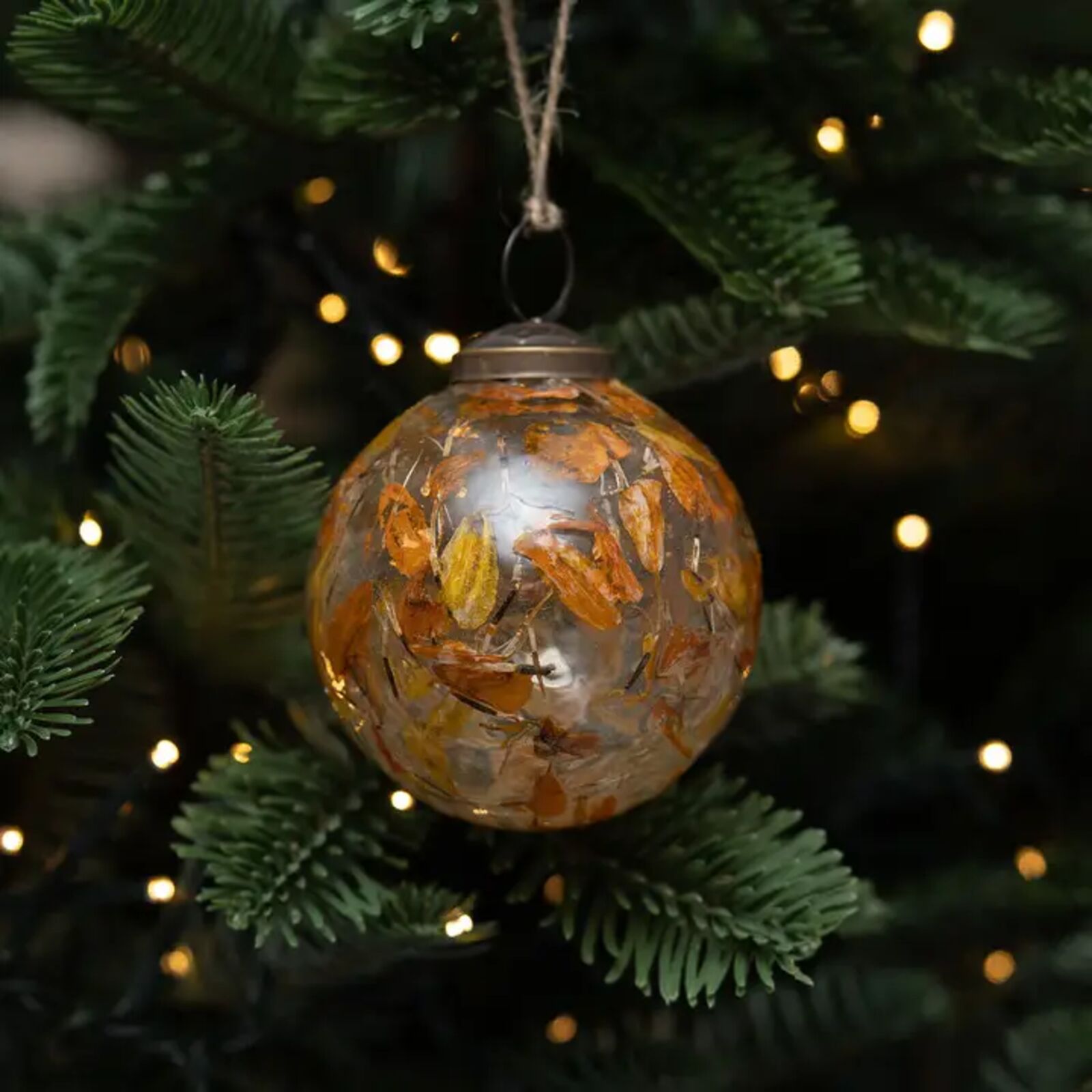 Decorative glass ornament with dried flowers hanging on a Christmas tree with lights.