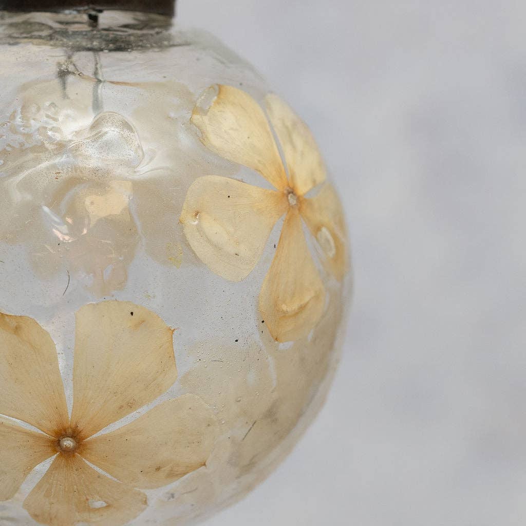 A close-up of a clear glass ornament or vase containing dried pale yellow flowers suspended in clear liquid, set against a soft light gray background.
