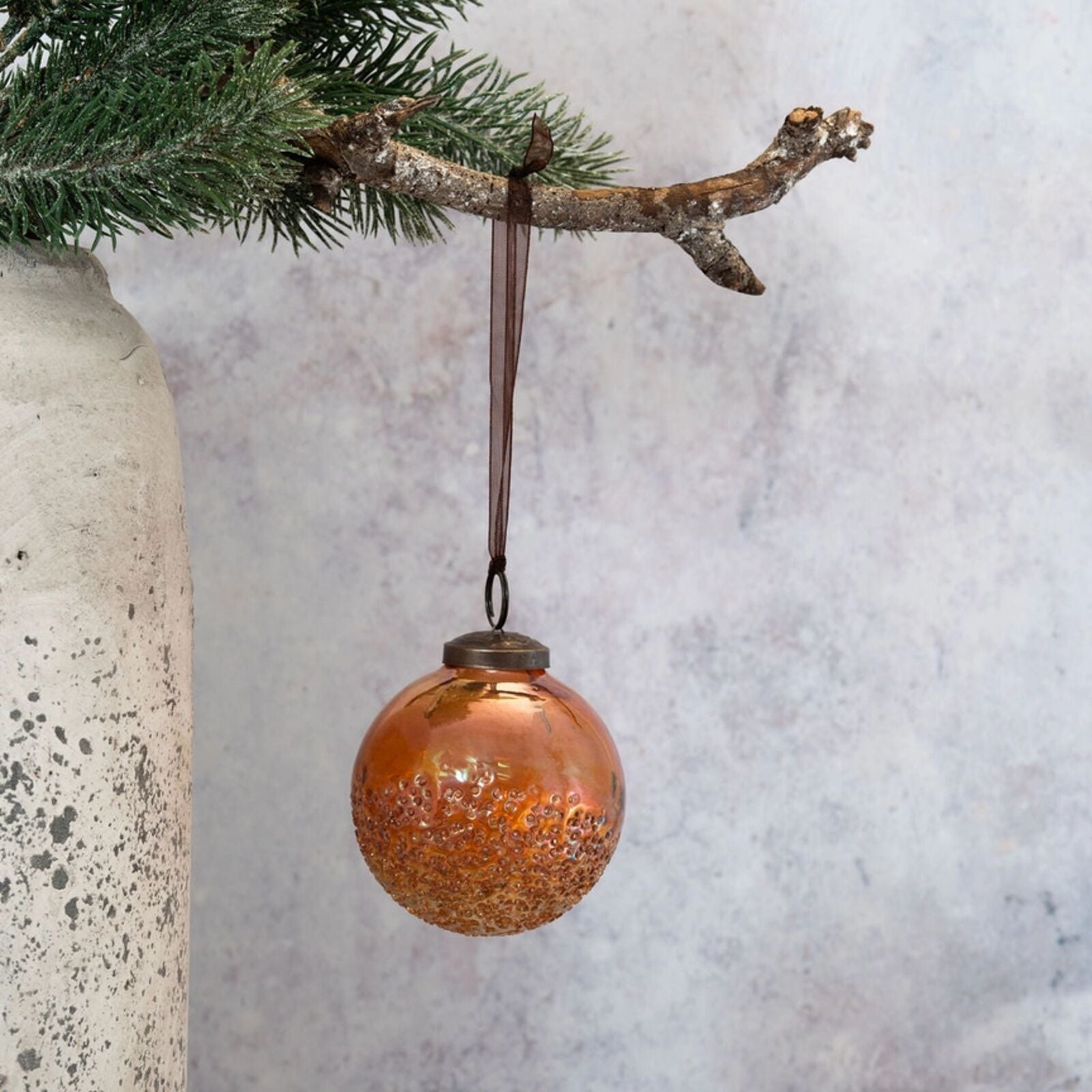 Bronze ornament hanging from a branch with a textured vase and light gray background