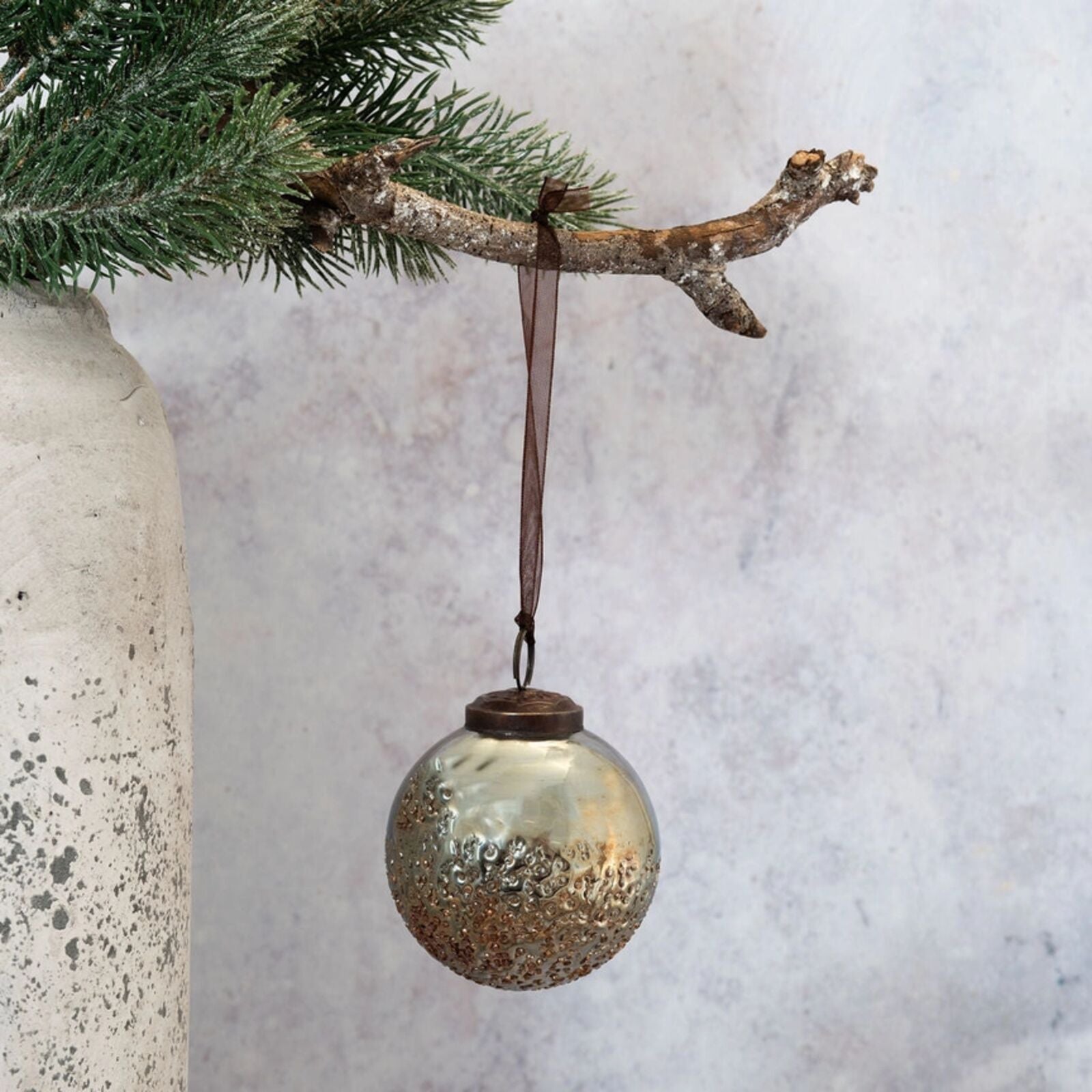 Decorative glass ball ornament hanging on a branch with a textured vase and white fabric background