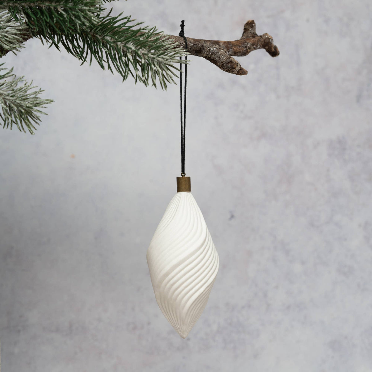 A white ribbed spiral Christmas ornament hanging from a black string with a brass cap, suspended from a snowy pine branch against a textured gray background.