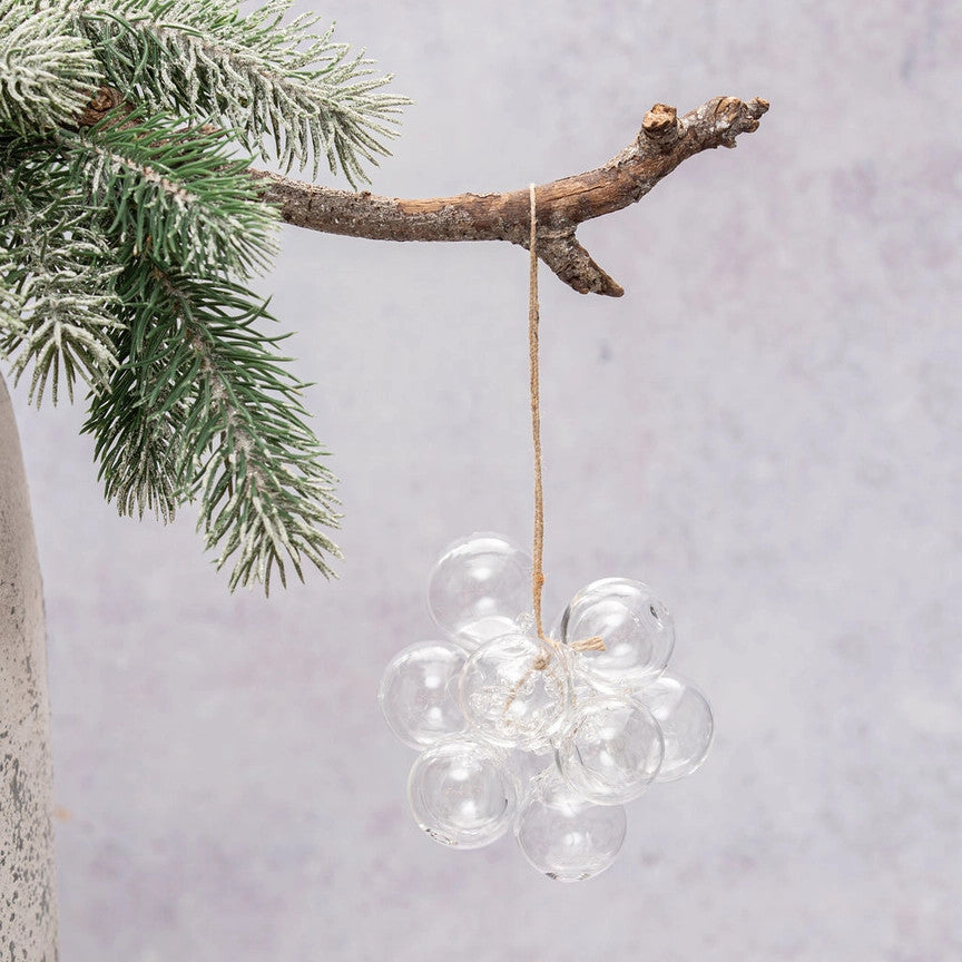 A cluster of clear glass bubble ornaments hanging from a natural twine string, suspended from a rustic tree branch with frosted green pine needles against a soft light gray background.