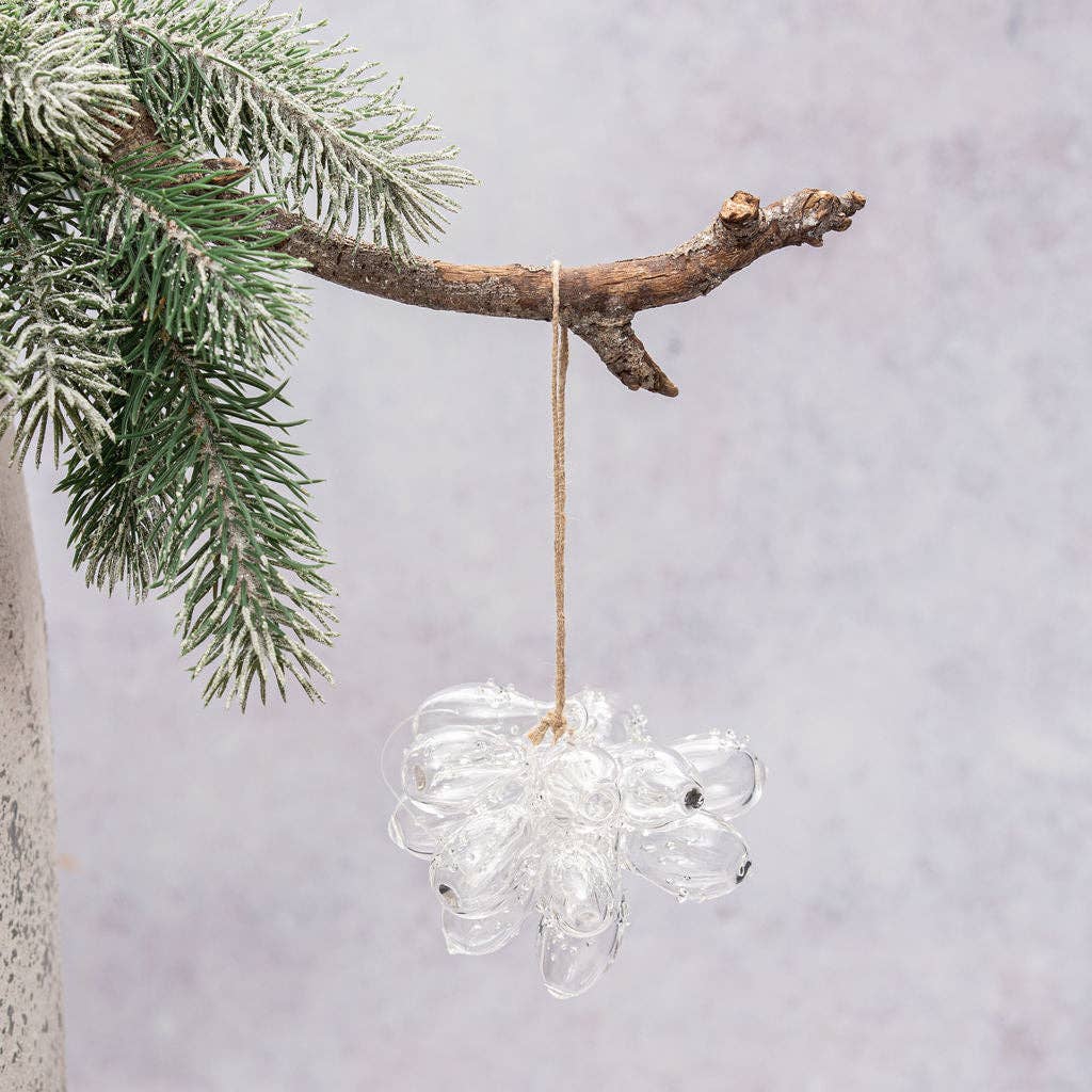 A clear glass hanging ornament shaped like a flower or cloud, suspended from a natural branch with green pine needles against a soft light gray background.