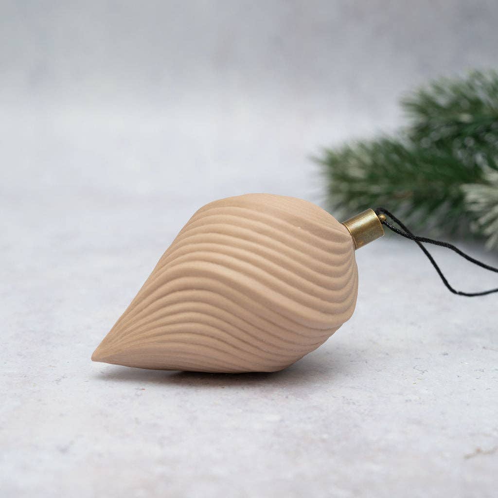 A natural wood Christmas ornament shaped like a teardrop or seashell with wavy grooved lines, featuring a brass hanging loop and black string, set against a light gray textured background with blurred green pine branches.