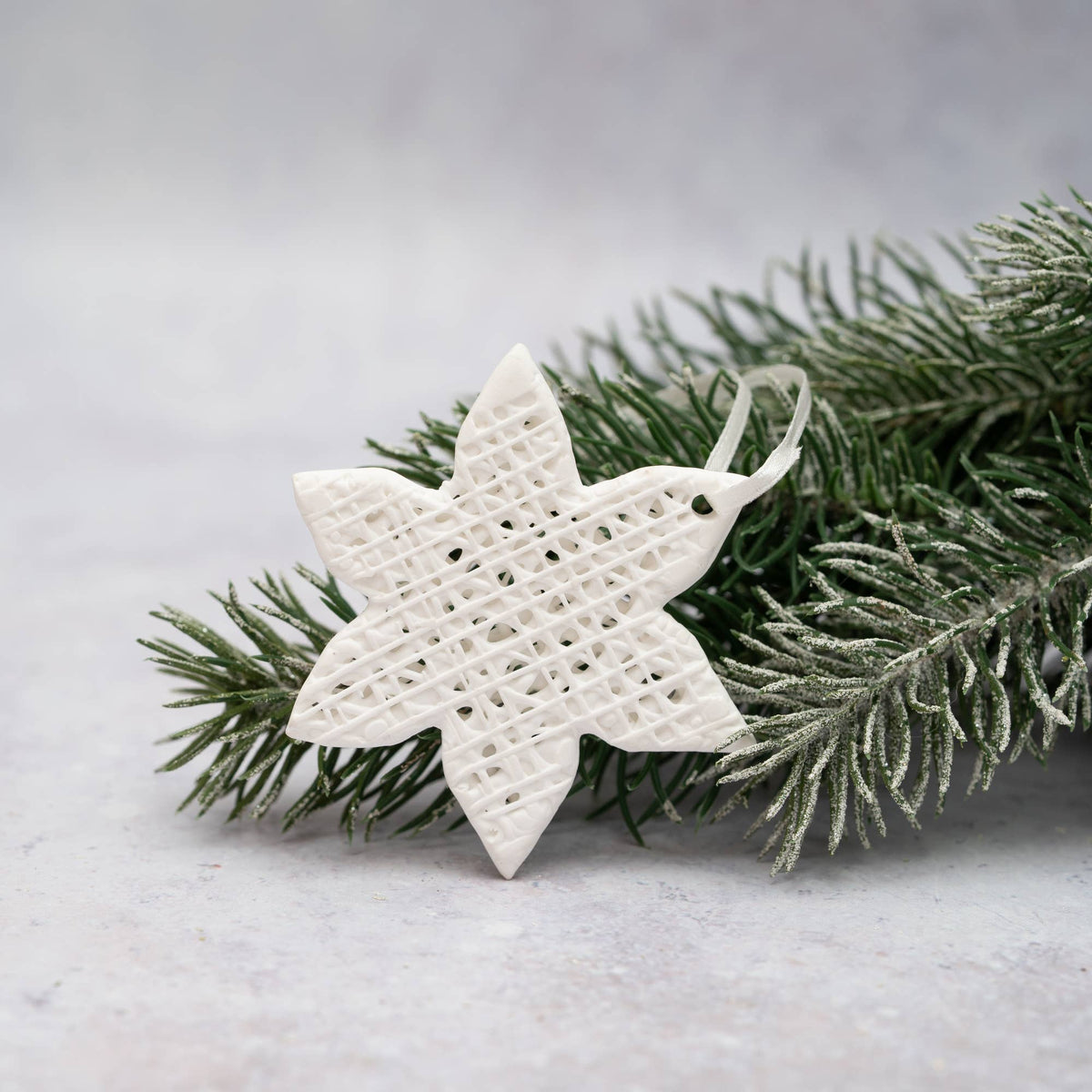 A white star-shaped hanging ornament with a woven basketweave pattern, displayed on green frosted pine branches against a light gray background.