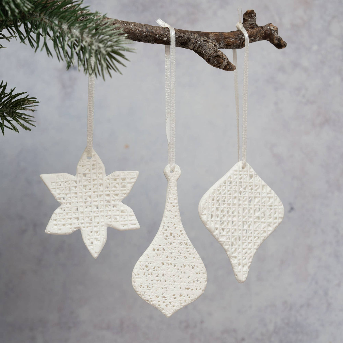 Three white textured ceramic Christmas ornaments shaped like a snowflake, a teardrop, and a diamond, hanging from a rustic tree branch with green pine needles against a soft gray background.