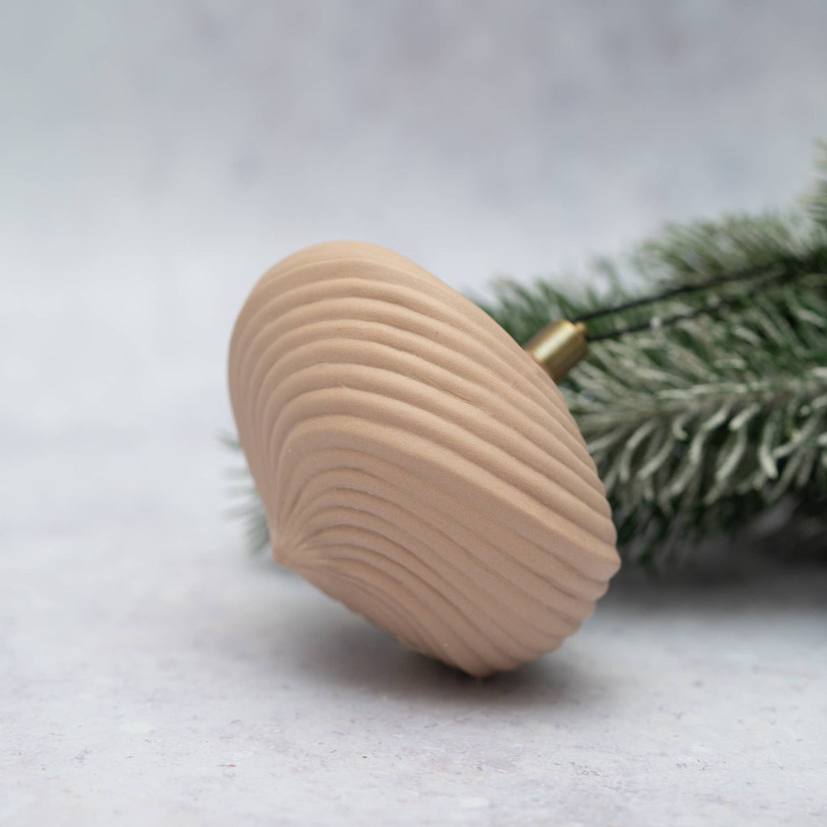 A natural wood Christmas dreidel with a ribbed shell-like pattern and a brass spindle, resting on a light gray surface with green pine needles in the background.