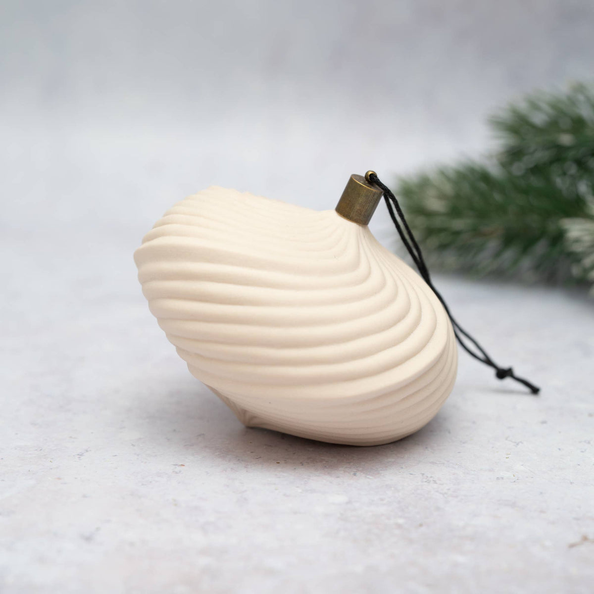 A cream-colored seashell-shaped ornament with a small brass grommet and black hanging string, resting on a light gray surface with blurred green pine branches in the background.