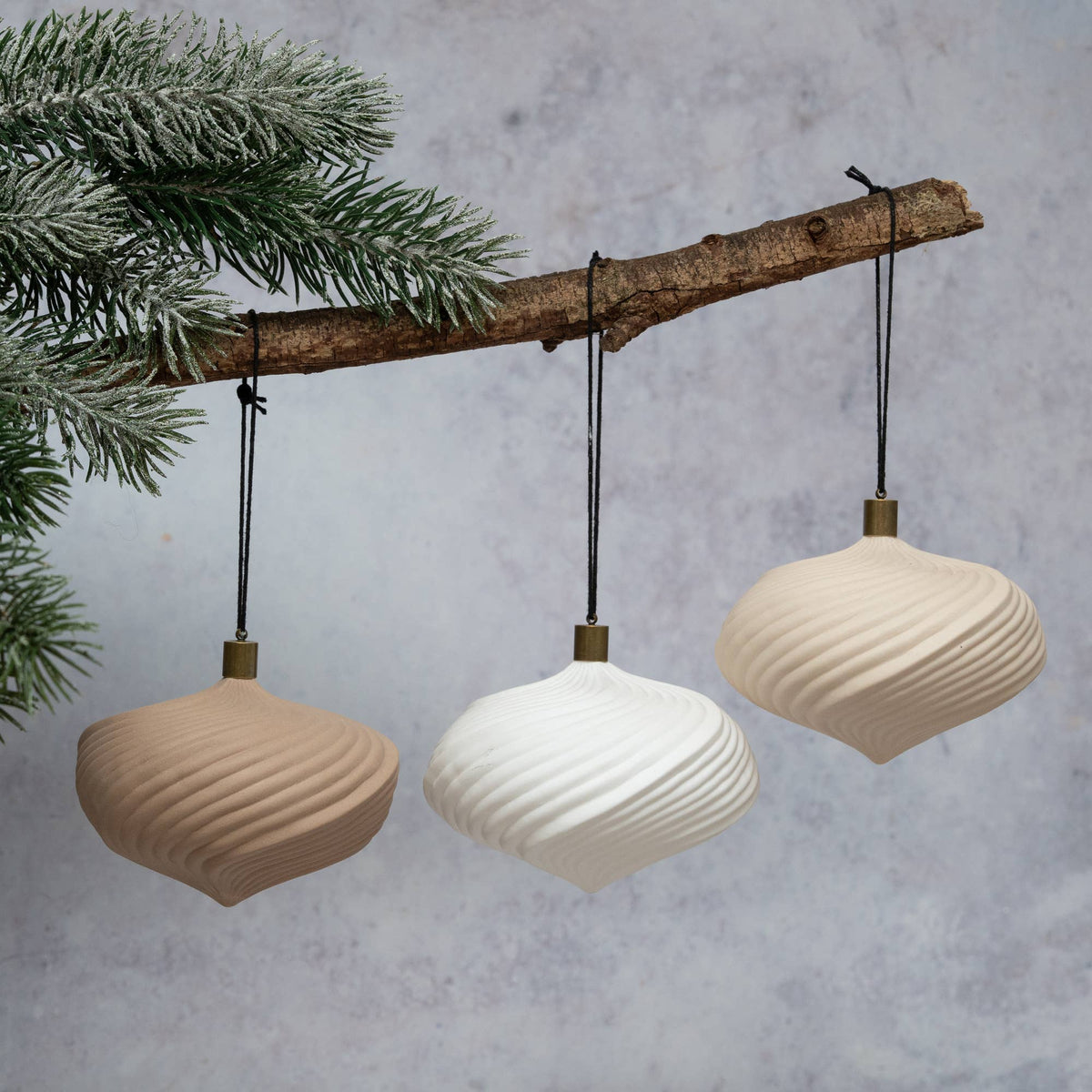 Three ribbed hanging pendant ornaments in beige, white, and cream, suspended from a rustic wooden branch with evergreen branches against a textured gray background.