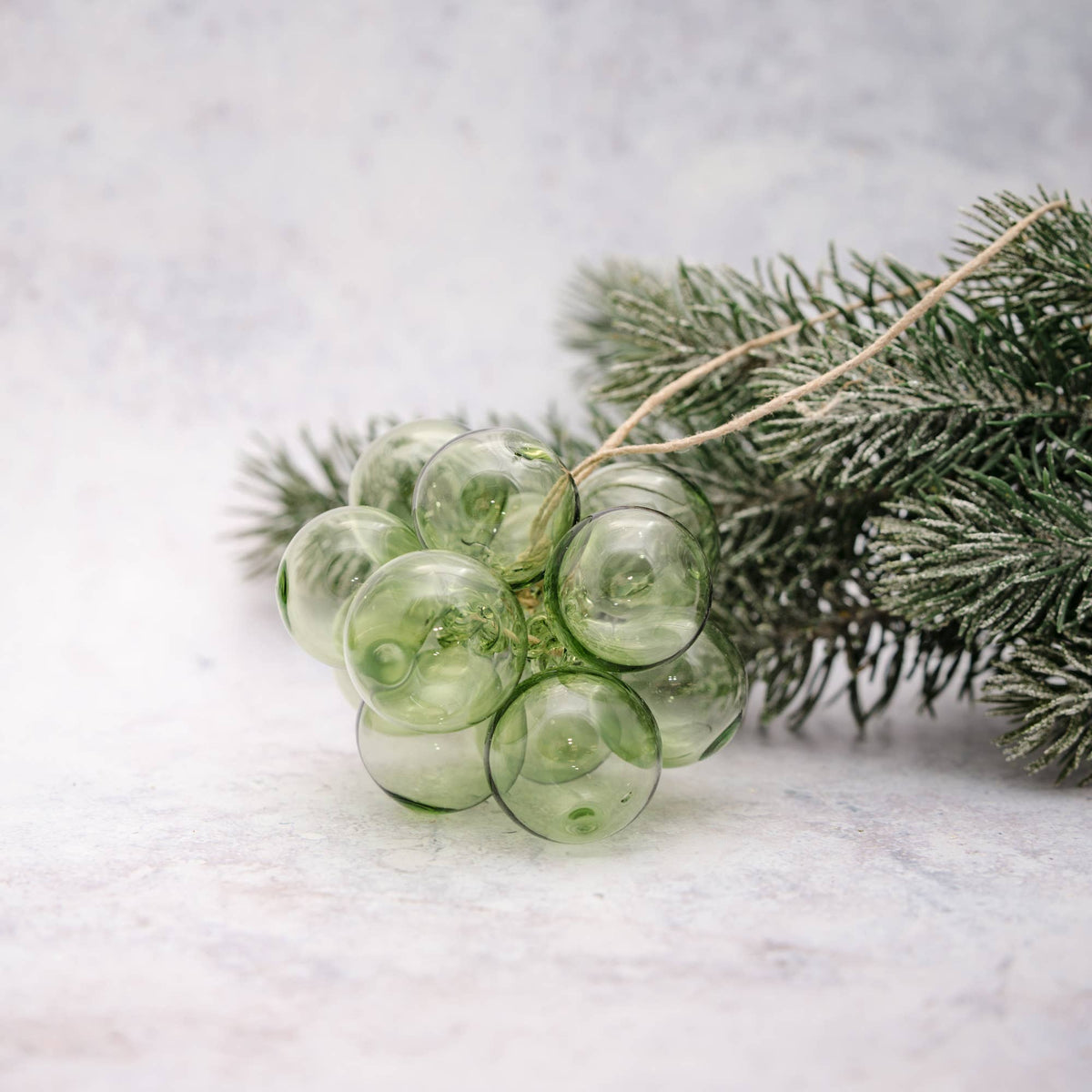 A cluster of translucent light green glass ornaments shaped like grapes, tied with a natural twine string and styled on a snowy pine branch against a light gray textured background.