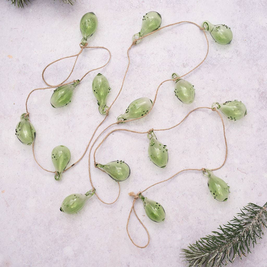 A garland of small, translucent light green glass ornaments hanging from a natural jute string, laid out on a pale textured surface with a pine branch in the corner.
