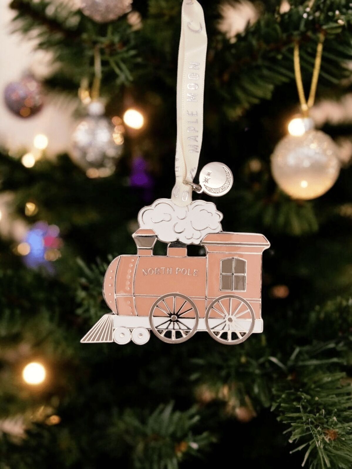 Decorative train ornament hanging on a Christmas tree with lights and ornaments in the background.