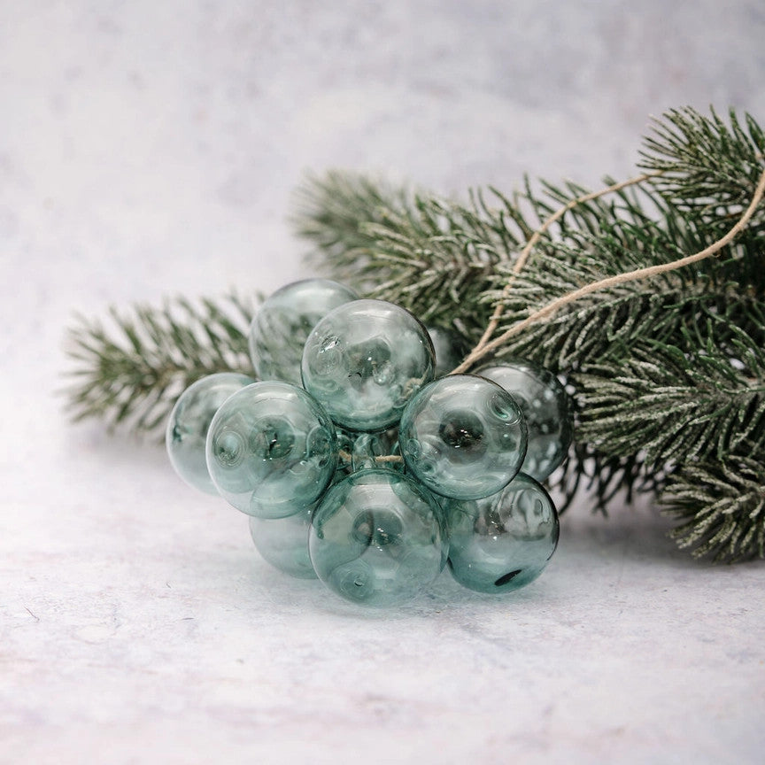 A cluster of translucent dusty blue-green glass ornaments shaped like pine berries, attached to a sprig of artificial evergreen branches on a light gray textured surface.
