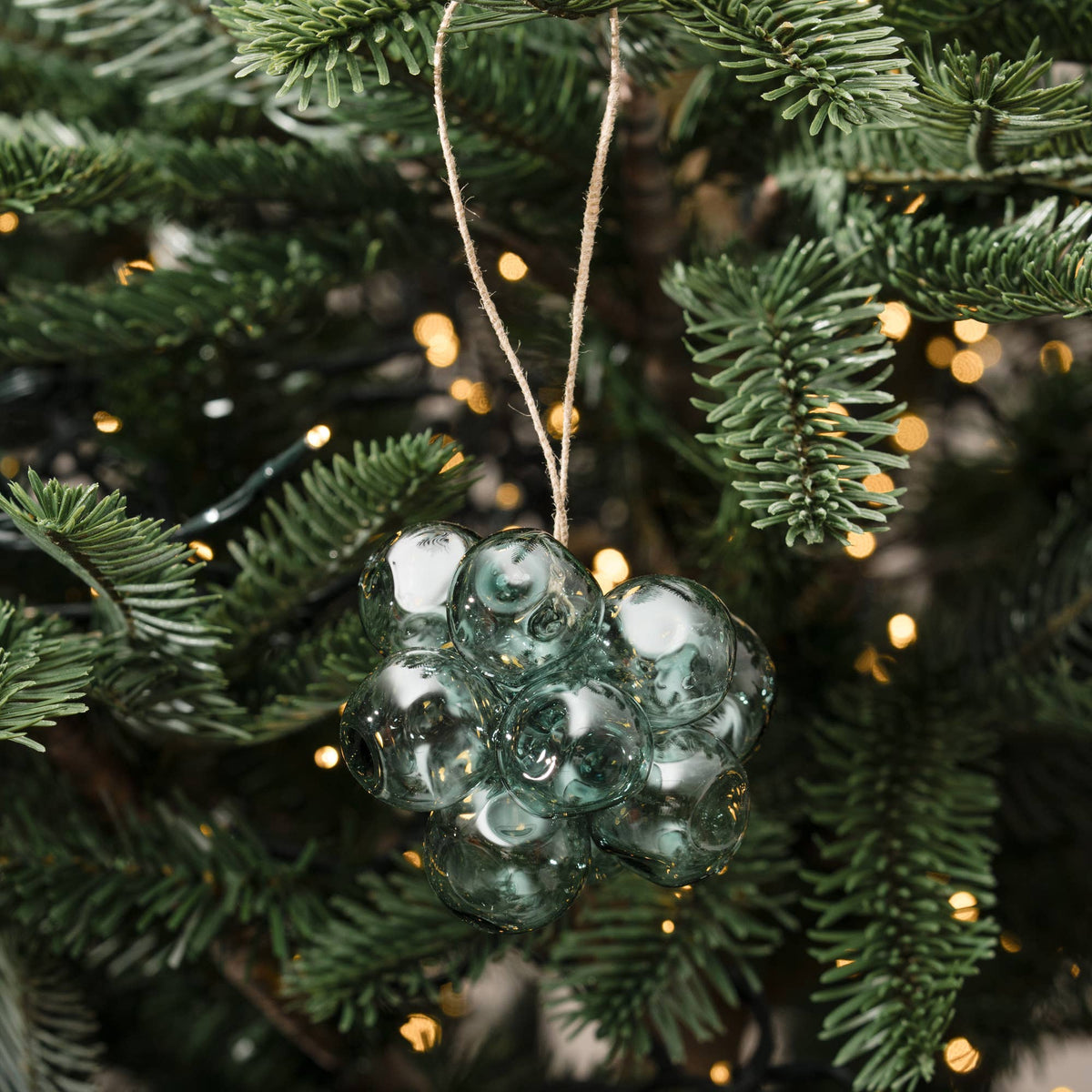 A green glass ornament shaped like a cluster of grapes hangs from a string on a pine tree with warm string lights in the background.