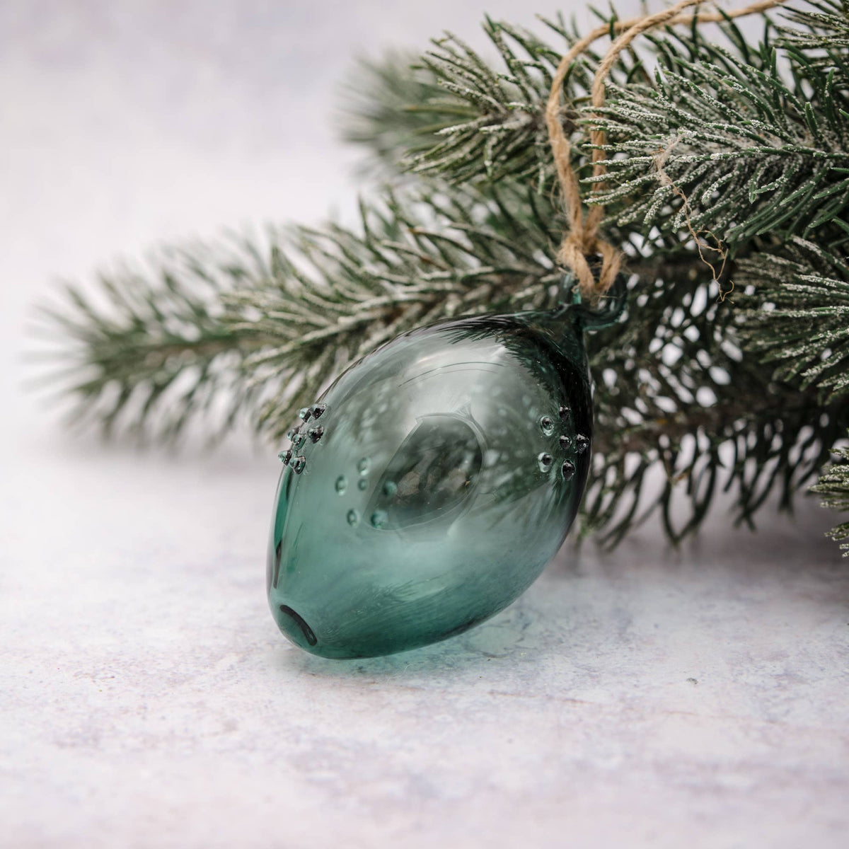 A translucent teal glass ornament hanging from a rustic twine string, nestled among frosted green pine branches against a soft, light gray background.