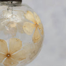 A close-up of a clear glass ornament or vase containing dried pale yellow flowers suspended in clear liquid, set against a soft light gray background.