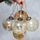 A cluster of clear glass Christmas ornaments decorated with pressed dried flowers and gold leaf, hanging from natural jute twine held by a hand with red nail polish.