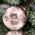 A close-up of a round, crackled glass Christmas ornament in a dusty rose or blush pink color, resting against green pine tree branches.