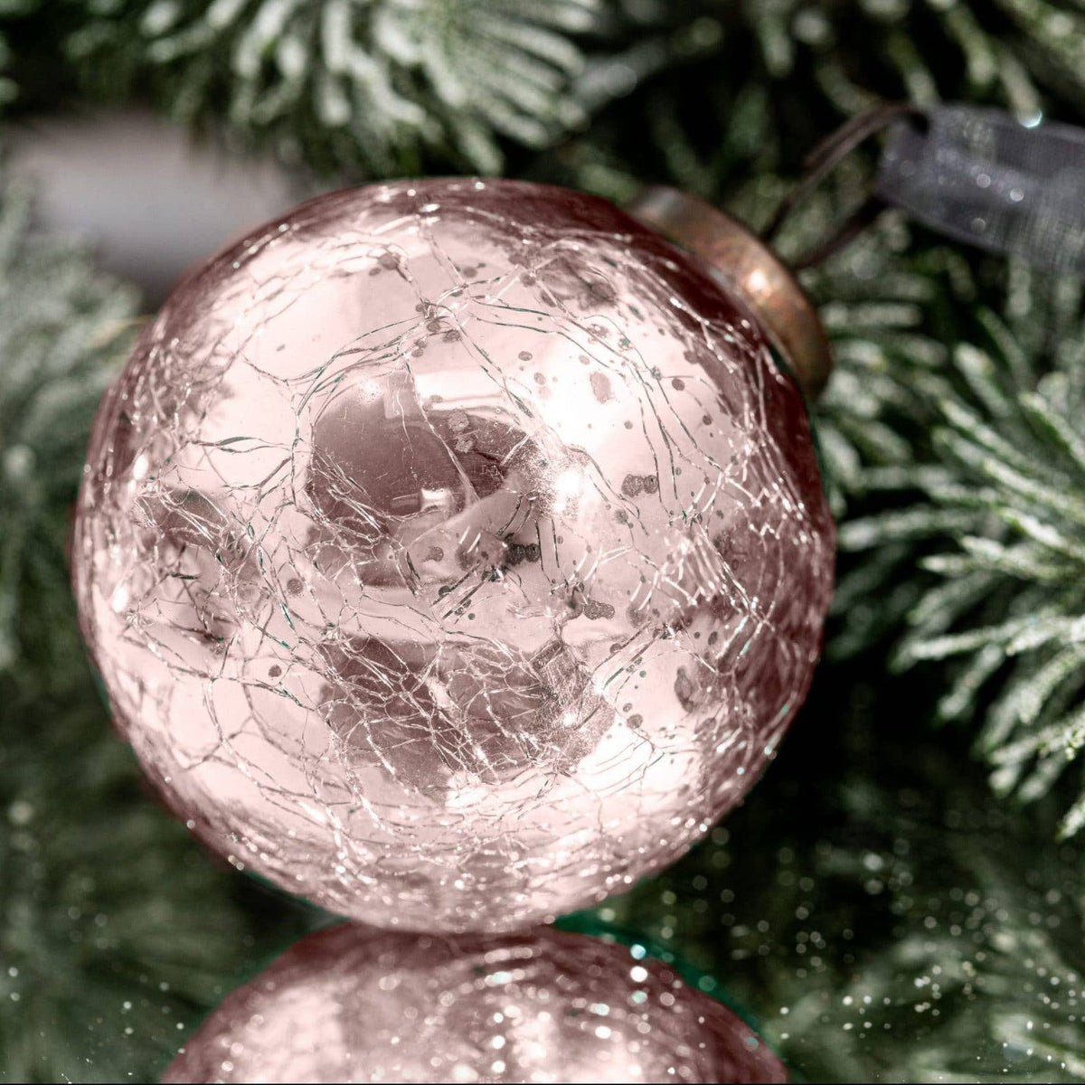 A close-up of a round, crackled glass Christmas ornament in a dusty rose or blush pink color, resting against green pine tree branches.