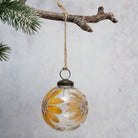 A clear glass Christmas ornament decorated with pressed yellow and brown daisy-like flowers, hanging from a natural twine loop attached to a rustic tree branch.