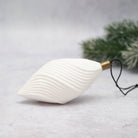 A white ribbed Christmas ornament shaped like a stylized leaf or teardrop, with a gold metal cap and black hanging string, photographed on a light gray surface with blurred green pine branches in the background.