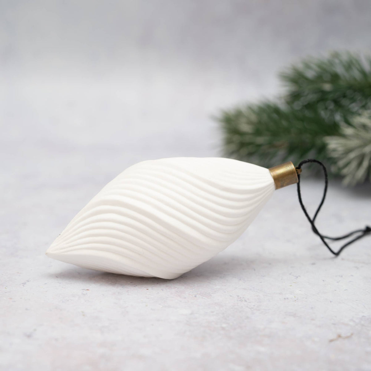 A white ribbed Christmas ornament shaped like a stylized leaf or teardrop, with a gold metal cap and black hanging string, photographed on a light gray surface with blurred green pine branches in the background.