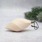 An off-white ceramic Christmas ornament shaped like a stylized leaf with wavy ridges, featuring a brass cap and a black hanging string, set against a light gray background with blurred green pine branches.