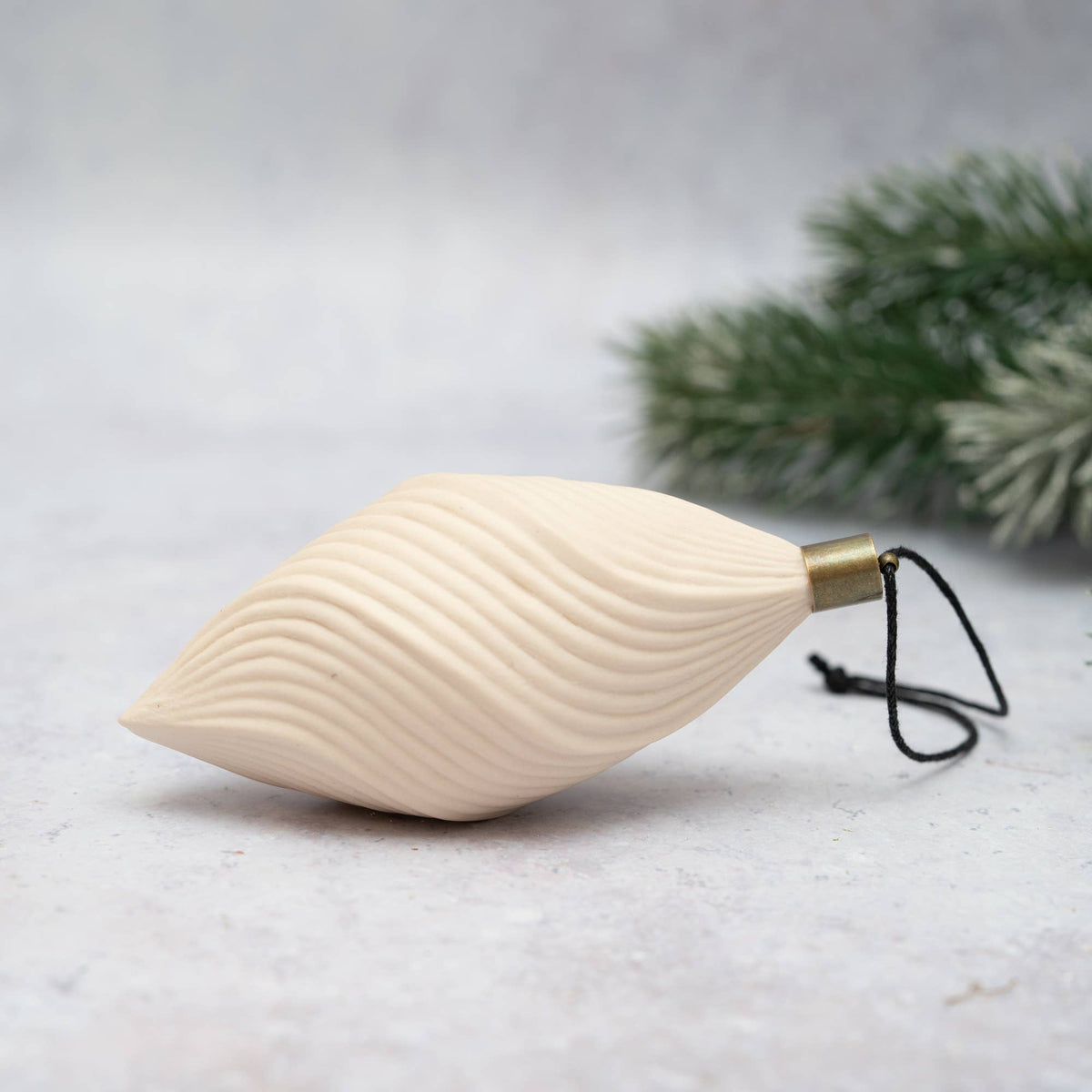 An off-white ceramic Christmas ornament shaped like a stylized leaf with wavy ridges, featuring a brass cap and a black hanging string, set against a light gray background with blurred green pine branches.