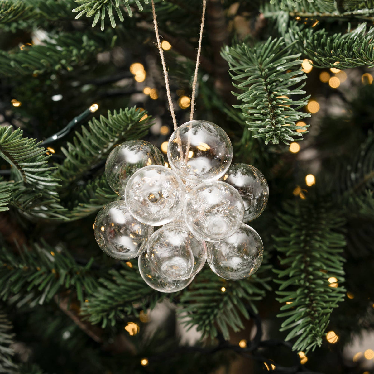 A cluster of clear glass Christmas ornaments shaped like a small flower or cluster of bubbles, hanging from a string on a green Christmas tree with warm string lights.