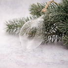 A clear glass teardrop-shaped Christmas ornament hanging from a natural twine loop attached to a frosted evergreen branch, set against a soft light gray background.