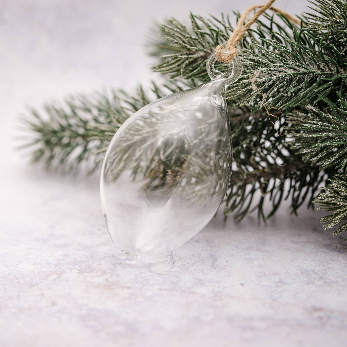 A clear glass teardrop-shaped Christmas ornament hanging from a natural twine loop attached to a frosted evergreen branch, set against a soft light gray background.