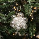 A cluster of clear glass Christmas ornaments shaped like a small flower or cluster of bubbles, hanging from a string on a green Christmas tree with warm string lights.