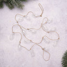 Clear glass teardrop ornaments hanging from a natural jute garland, arranged on a light textured surface with pine branches in the corners.