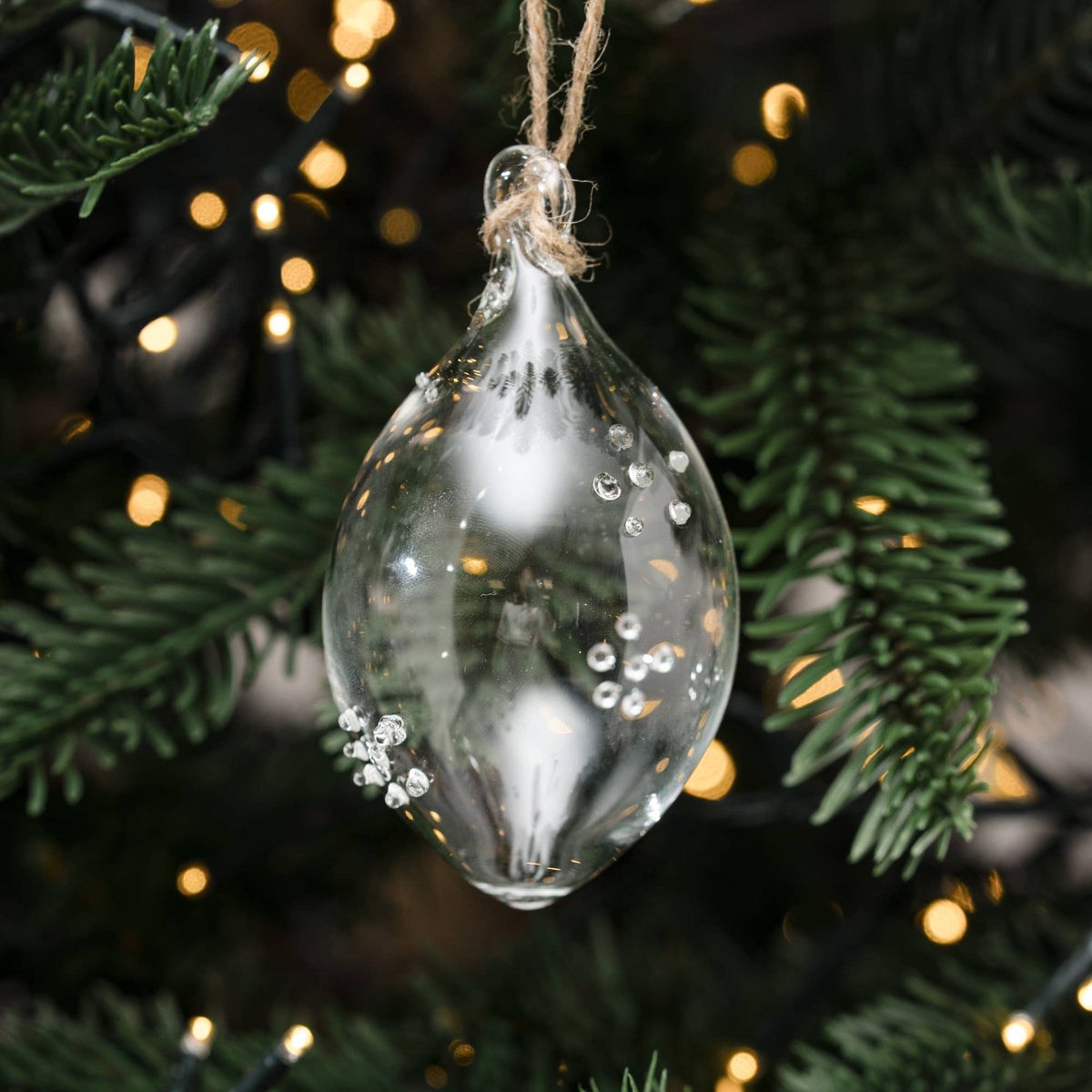 A clear glass Christmas ornament shaped like a teardrop, hanging from a jute string on a decorated Christmas tree with warm lights and green pine branches.
