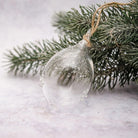A clear glass Christmas ornament with delicate snowflake patterns and a natural jute hanging string, resting on green frosted pine branches against a soft, light gray background.