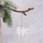 A clear glass hanging ornament shaped like a flower or cloud, suspended from a natural branch with green pine needles against a soft light gray background.