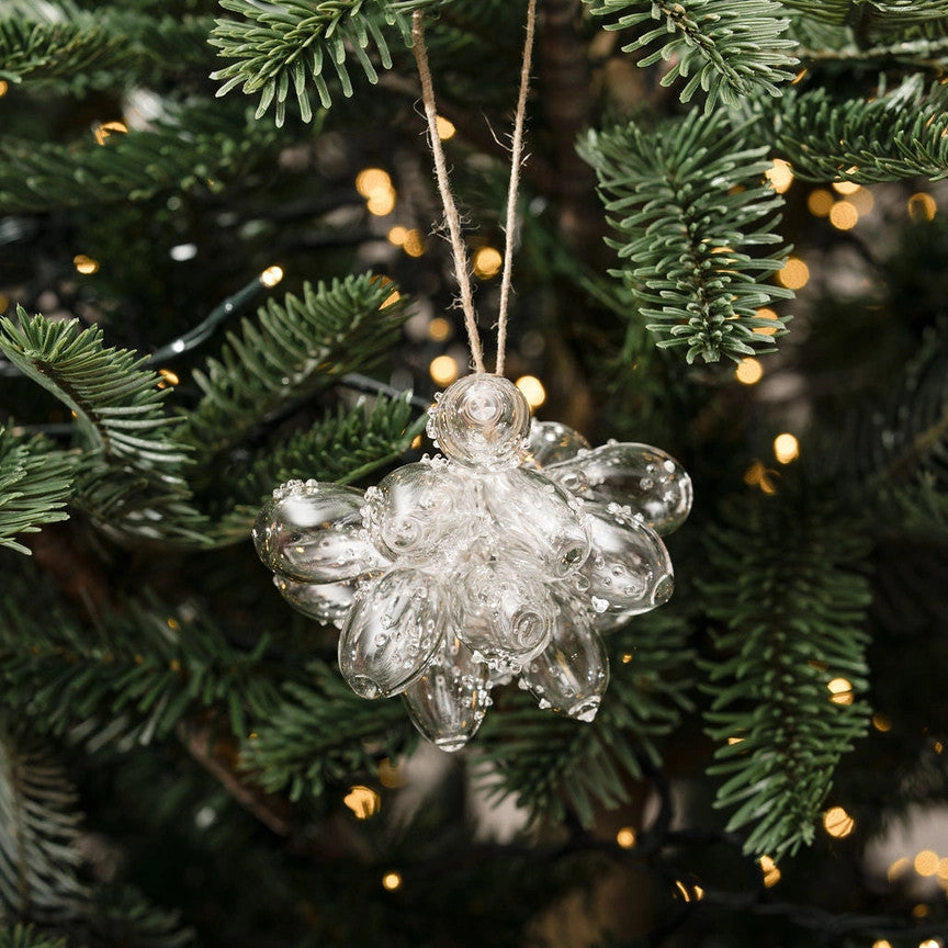 A clear glass Christmas ornament shaped like a flower or snowflake hangs from a twine string among green pine tree branches with warm yellow bokeh lights in the background.