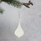 A white textured ceramic Christmas ornament shaped like a teardrop, hanging from a pine branch against a light gray background.