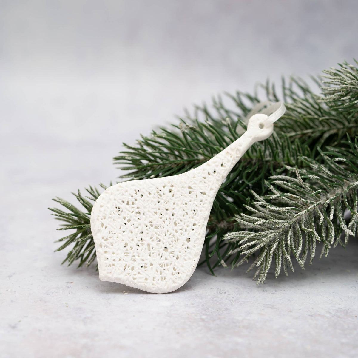 A white ceramic Christmas ornament shaped like a pear with an intricate openwork lattice pattern, resting on a sprig of frosted green pine branches against a light gray background.