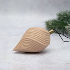A natural wood Christmas ornament shaped like a teardrop or seashell with wavy grooved lines, featuring a brass hanging loop and black string, set against a light gray textured background with blurred green pine branches.