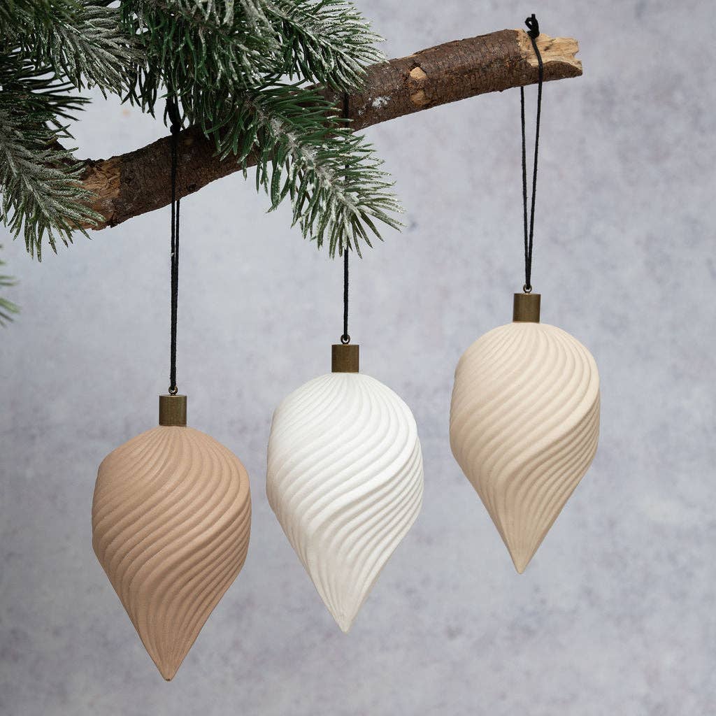 Three ribbed ceramic Christmas tree ornaments in beige, white, and cream hanging from a pine branch against a gray background.