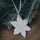 A white star-shaped ceramic Christmas ornament with a woven lattice pattern, hanging from a white ribbon against a background of green pine branches and a wooden surface.