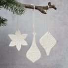Three white textured ceramic Christmas ornaments shaped like a snowflake, a teardrop, and a diamond, hanging from a rustic tree branch with green pine needles against a soft gray background.