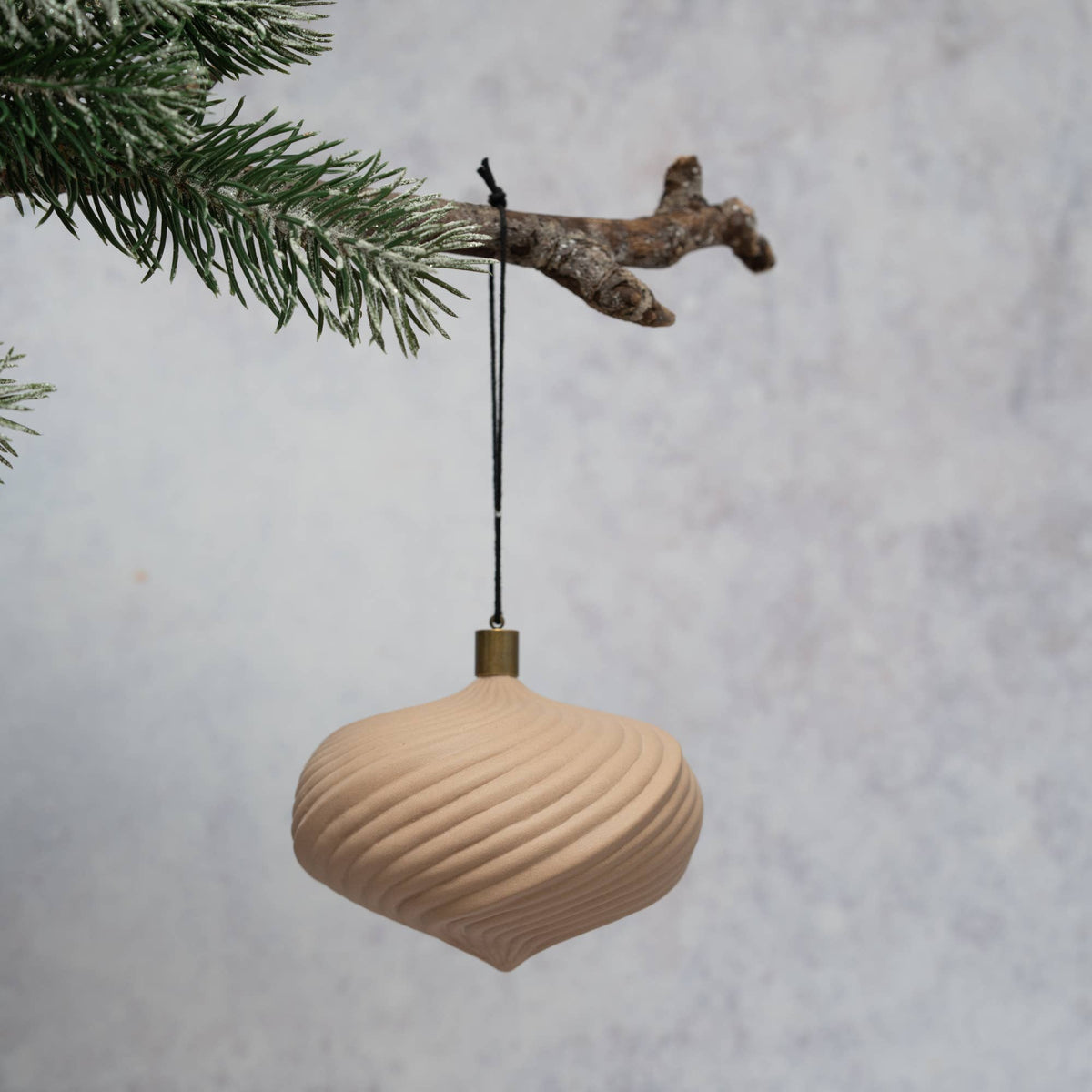 A ribbed, teardrop-shaped wooden Christmas ornament in a light natural beige finish, hanging from a rustic tree branch with a black hanging string against a textured gray background.