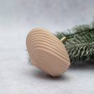 A natural wood Christmas dreidel with a ribbed shell-like pattern and a brass spindle, resting on a light gray surface with green pine needles in the background.