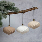 Three ribbed hanging pendant ornaments in beige, white, and cream, suspended from a rustic wooden branch with evergreen branches against a textured gray background.