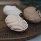 Three shell-shaped ceramic ornaments in white, beige, and taupe, arranged on a dark wooden tray with a shallow depth of field.