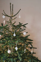 A green Christmas tree decorated with white hanging ornaments and warm white string lights against a plain beige wall.