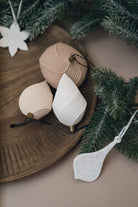 Three ribbed ceramic Christmas tree ornaments in beige, cream, and white, styled on a wooden tray with green pine branches and a white hanging ornament against a neutral background.