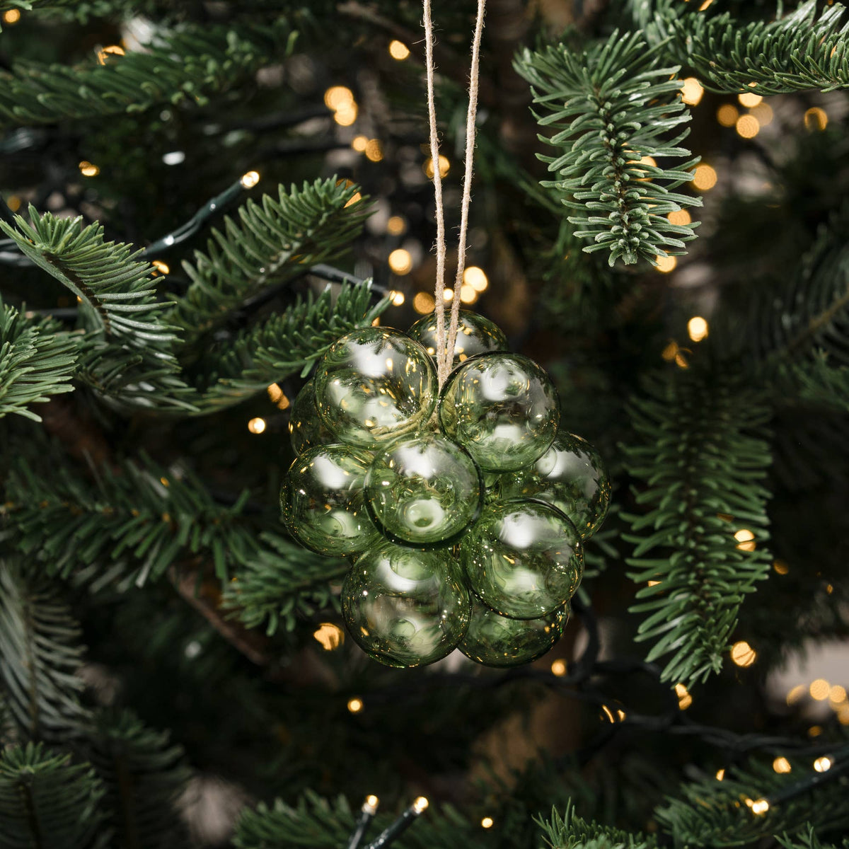 A green glass ornament shaped like a cluster of grapes hangs on a Christmas tree with green needles and warm white string lights in the background.